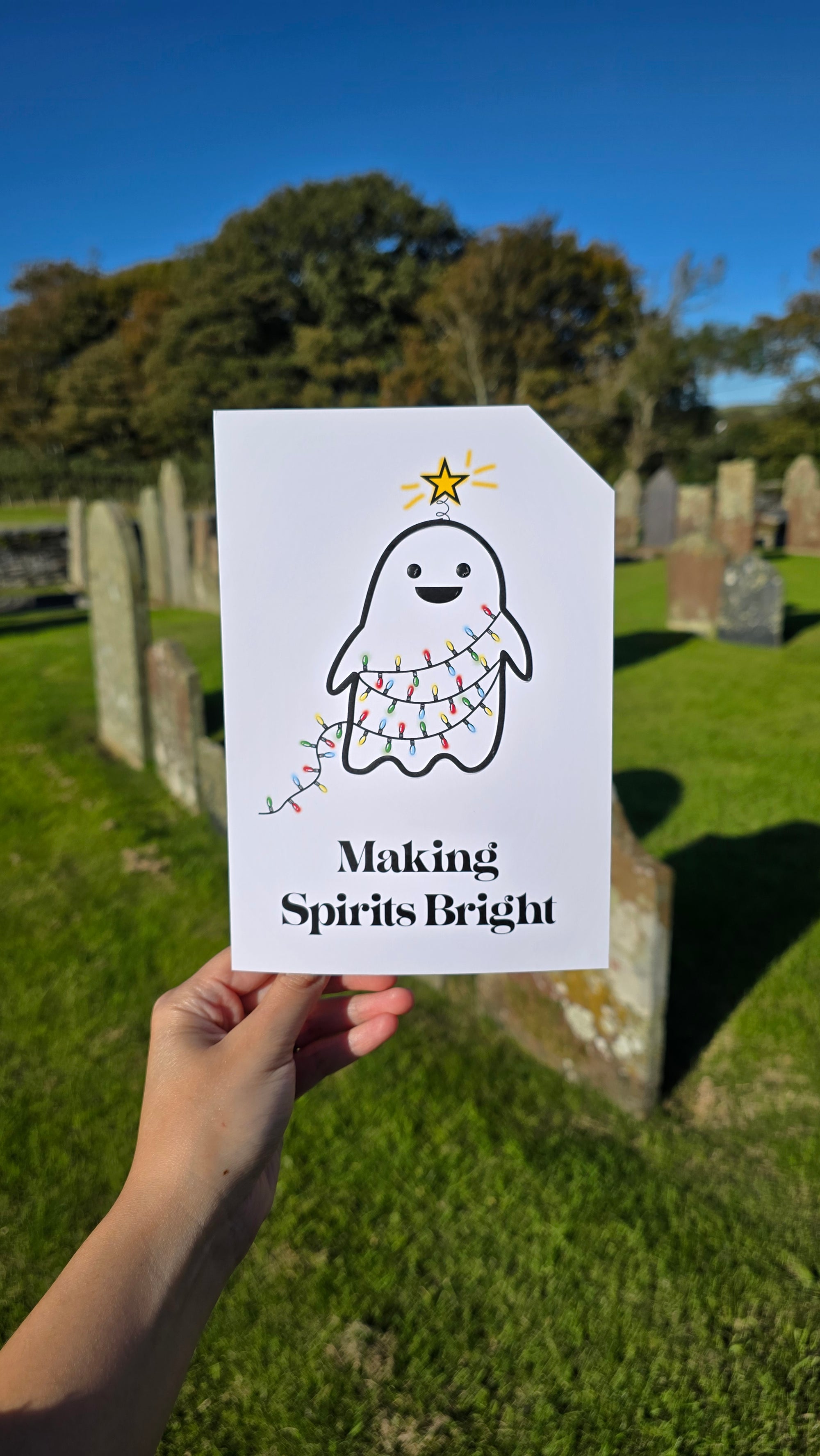 Card front with a smiling ghost wrapped in red, yellow, green and blue, Christmas tree lights. The ghost has a glowing yellow star on its head.
The text on the bottom reads 'Making Spirits Bright', in bold black letters. The top right hand corner is cut off, for orientation purposes; a unique Dot & Type feature.
The image was taken in a graveyard, the card is in the foreground and there are old graves in the background. The sky is bright blue and cloudless.