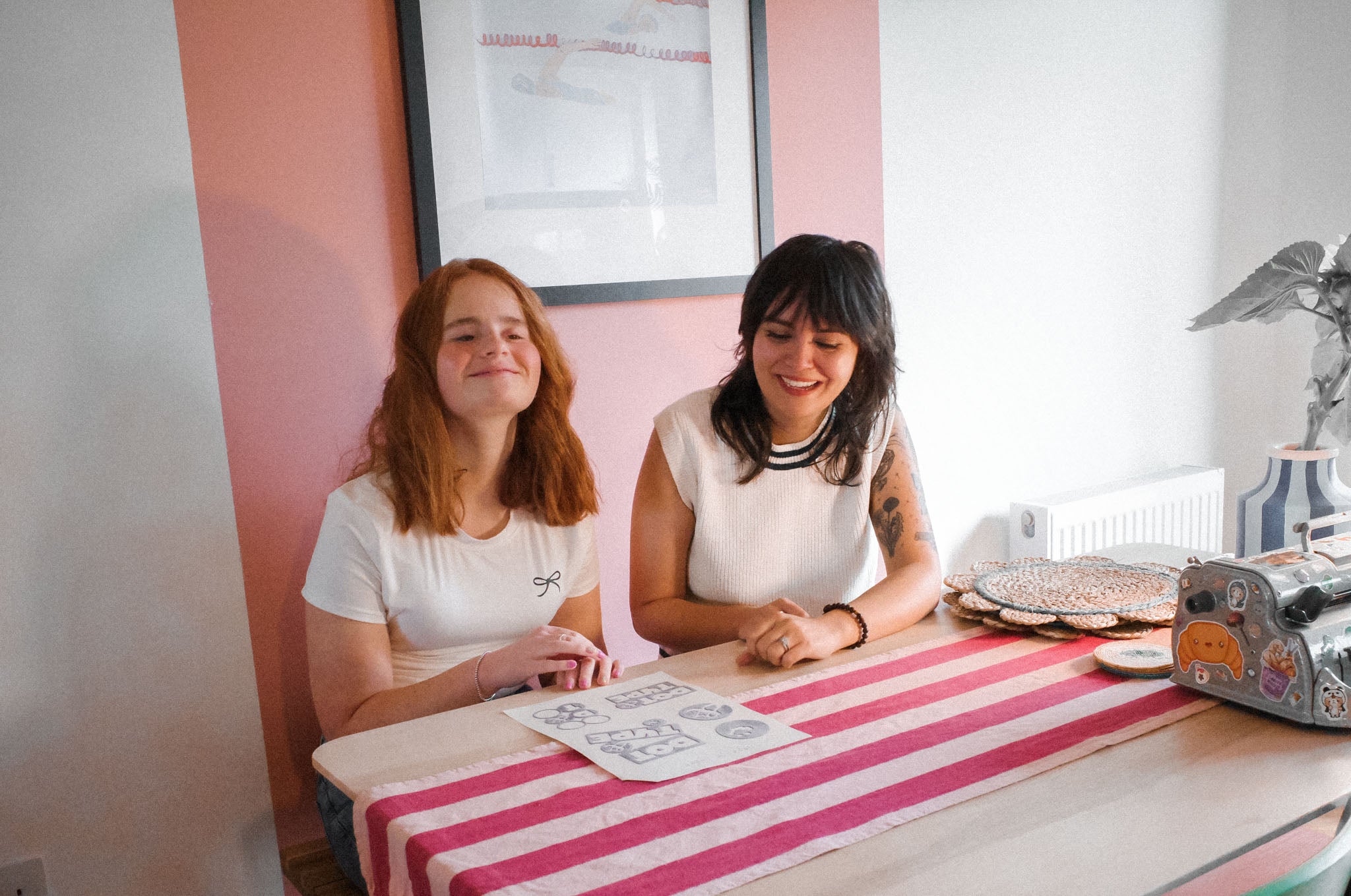 Two women (one with red hair and a visual impairment and one with dark hair) sitting at a table with a pink and white striped tablecloth underneath a tactile image in front of them, both smiling.