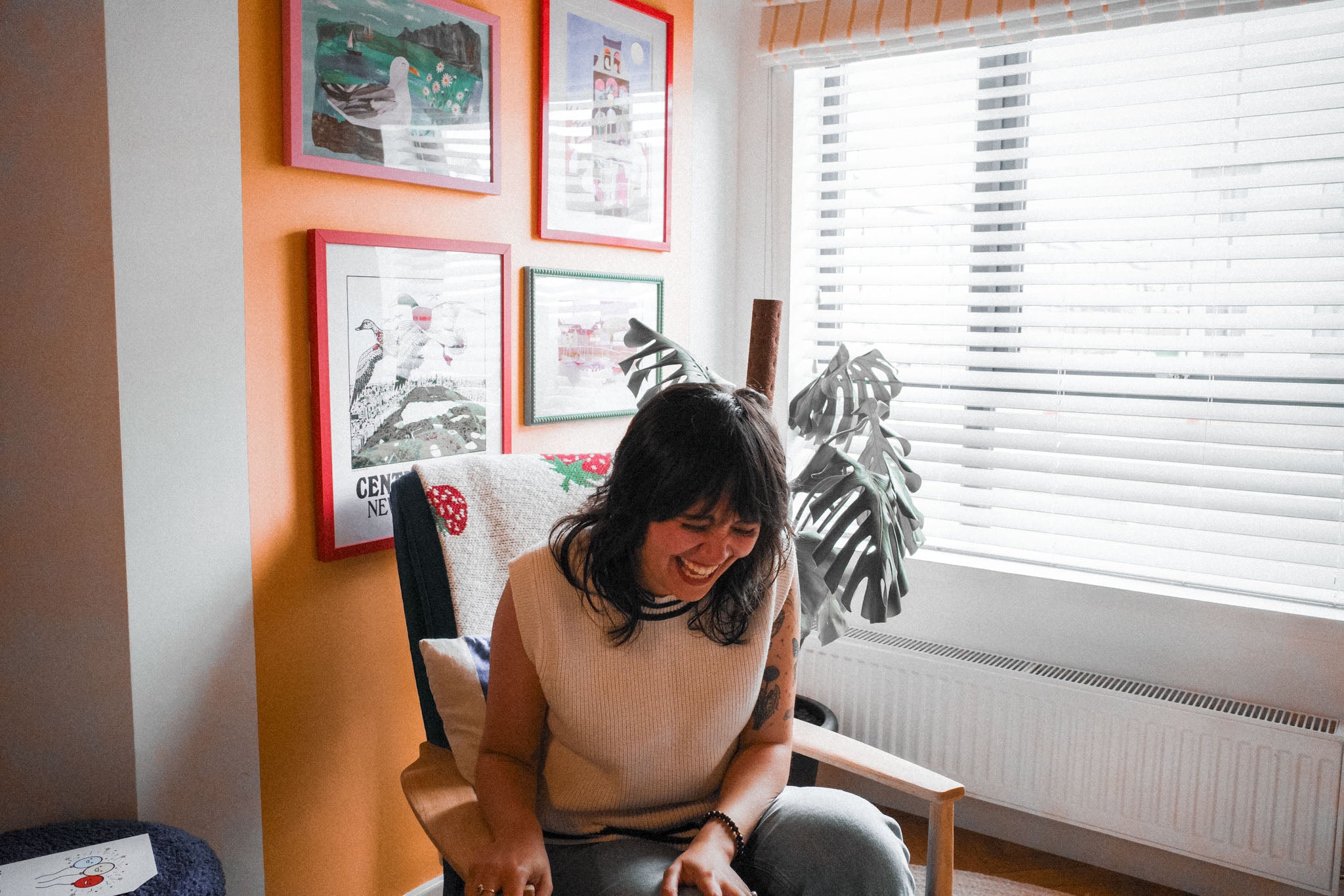 Woman sitting in a room with framed pictures on the wall. Laughing, clutching the right arm of the armchair.
