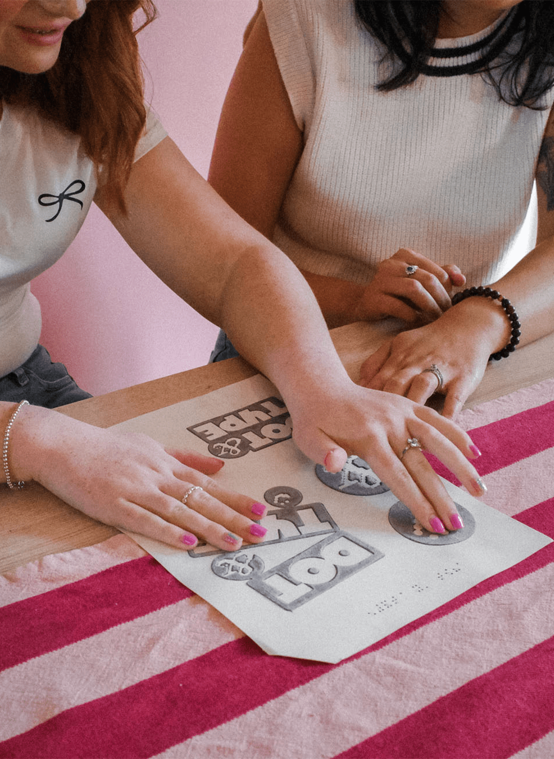 Two people sat next to each other at a table. There is a blind woman who is reading a tactile diagram on the table. It is covered in Dot & Type logos.