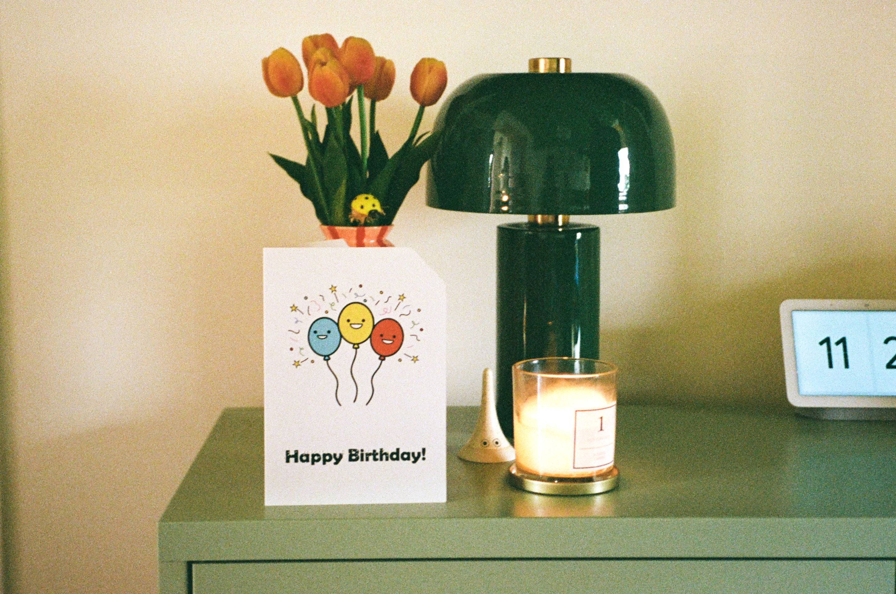 Green lamp on a table with a birthday card, flowers, and a lit candle. The card has 3 colourful balloons on the front with its top right-hand corner cut off.