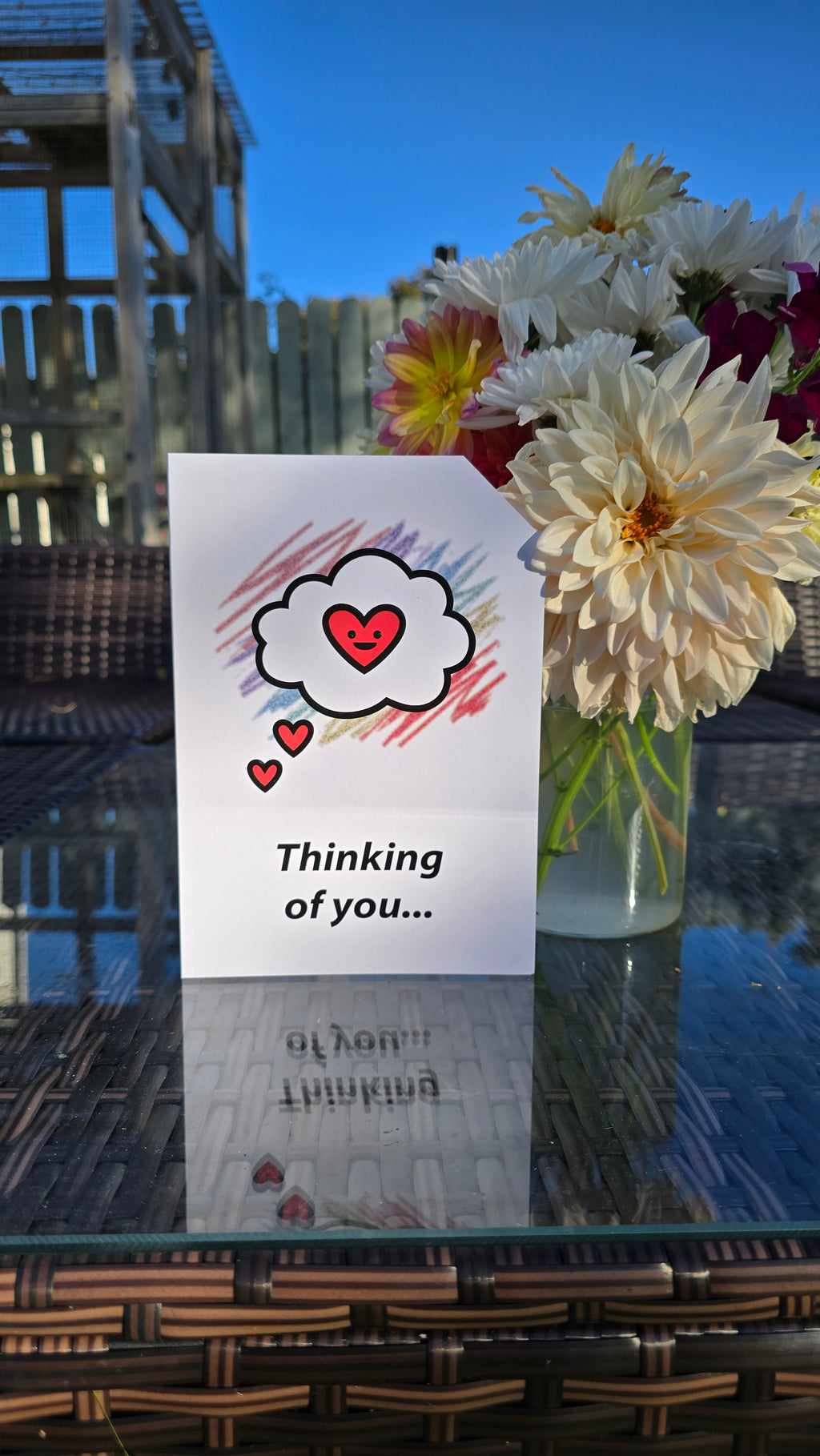 Card front with a 'thinking cloud' with a smiling red heart in the centre, in the foreground. Colourful aray of scribbles in the background.
The text on the bottom reads 'Thinking of you...', in bold black letters. The top right hand corner is cut off, for orientation purposes; a unique Dot & Type feature.
The image shows the card on a table with a small vase of flowers next to it. There are large homegrown dahlias in the vase.