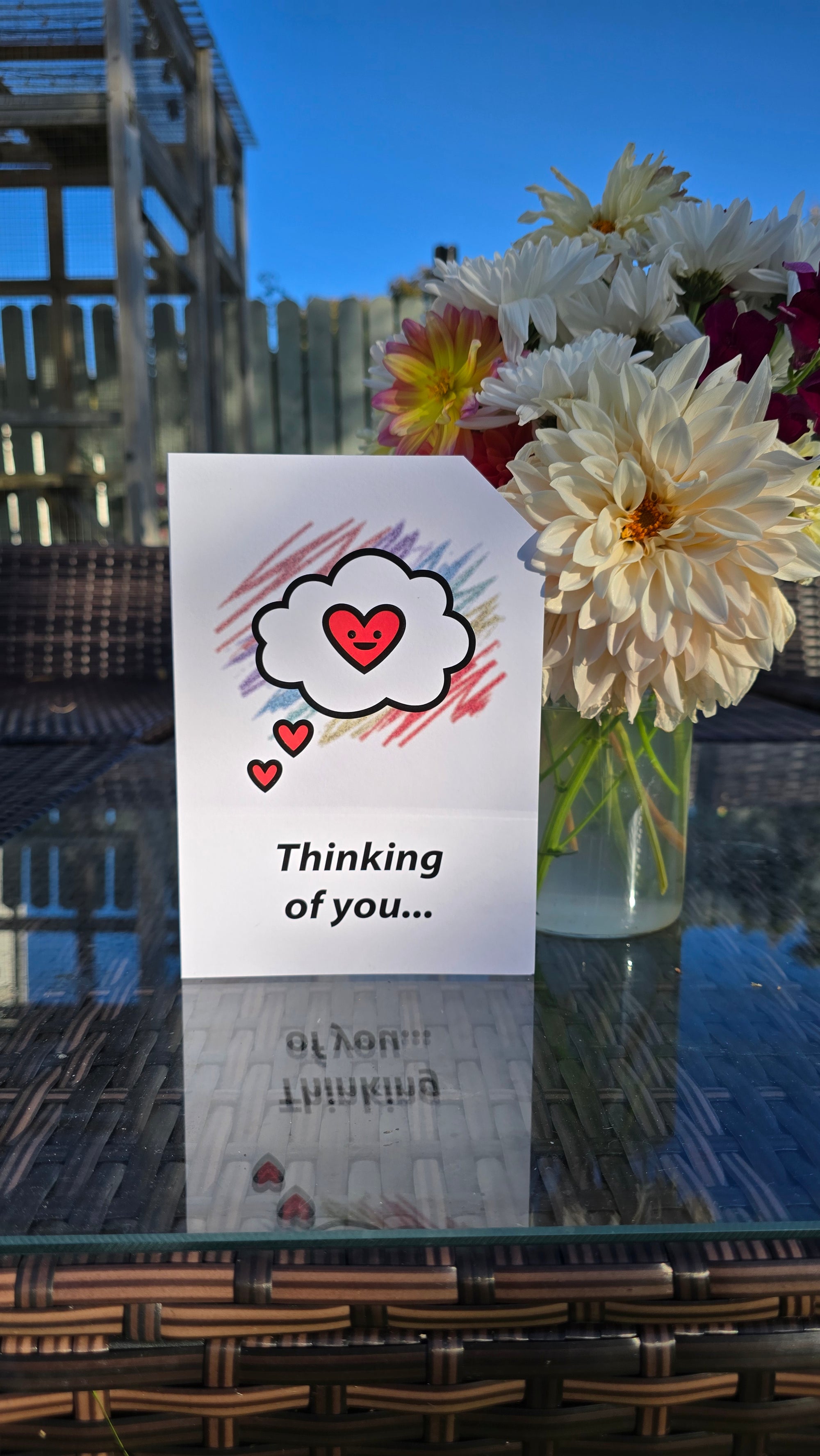 Card front with a 'thinking cloud' with a smiling red heart in the centre, in the foreground. Colourful aray of scribbles in the background.
The text on the bottom reads 'Thinking of you...', in bold black letters. The top right hand corner is cut off, for orientation purposes; a unique Dot & Type feature.
The image shows the card on a table with a small vase of flowers next to it. There are large homegrown dahlias in the vase.