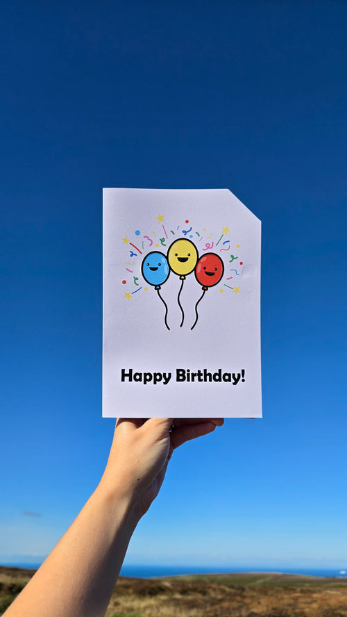 Card front with 3 smiling, cartoon balloons, with bright confetti type graphics in the background. Left balloon is pale blue, middle balloon is yellow and the right balloon is red.
The text on the bottom reads 'Happy Birthday!', in bold black letters. The top right hand corner is cut off, for orientation purposes; a unique Dot & Type feature.
The image is of the card being held in front of a bright blue, cloudless sky with the landscape at the very bottom of the screen.