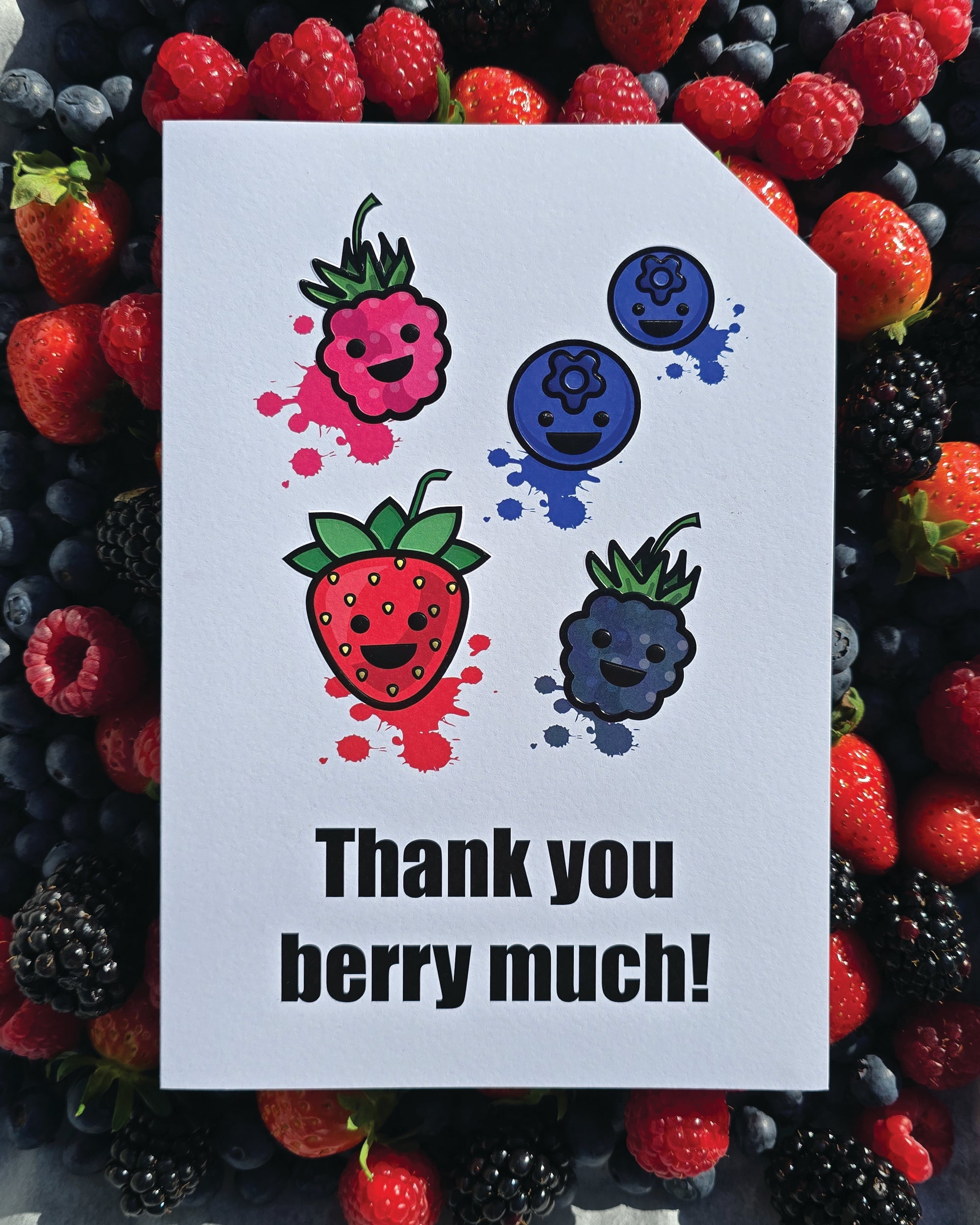 Card front with various smiling berries in the foreground (1 raspberry, 1 strawberry, 1 blackberry and 2 blueberries). Colourful splats in the background.
The text on the bottom reads 'Thank you berry much!', in bold black letters. The top right hand corner is cut off, for orientation purposes; a unique Dot & Type feature.
The image is of the card placed on top of a series of berries which match the card front. They are laid on a baking tray.