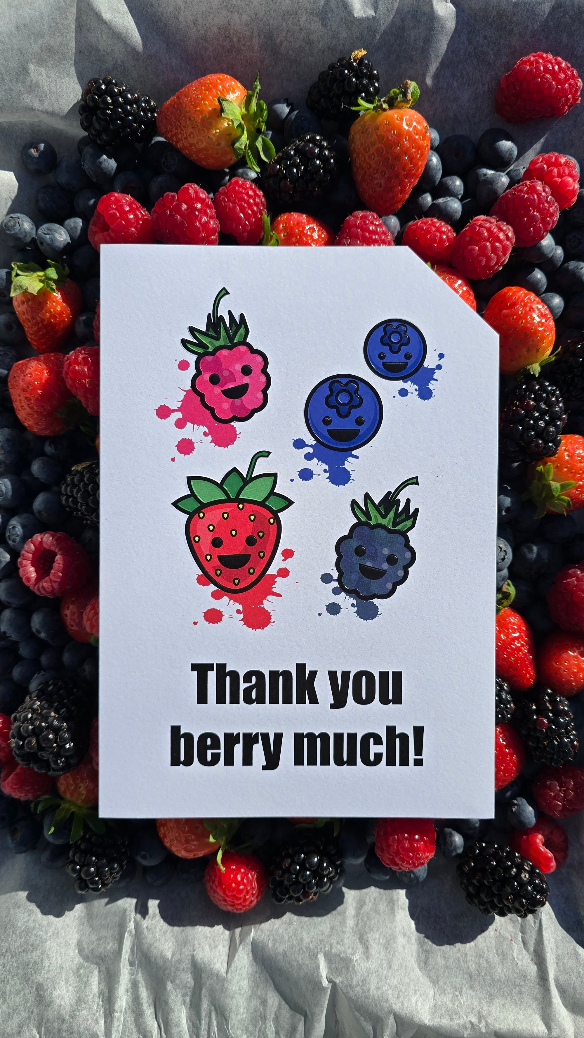 Card front with various smiling berries in the foreground (1 raspberry, 1 strawberry, 1 blackberry and 2 blueberries). Colourful splats in the background.
The text on the bottom reads 'Thank you berry much!', in bold black letters. The top right hand corner is cut off, for orientation purposes; a unique Dot & Type feature.
The image is of the card placed on top of a series of berries which match the card front. They are laid on a baking tray.