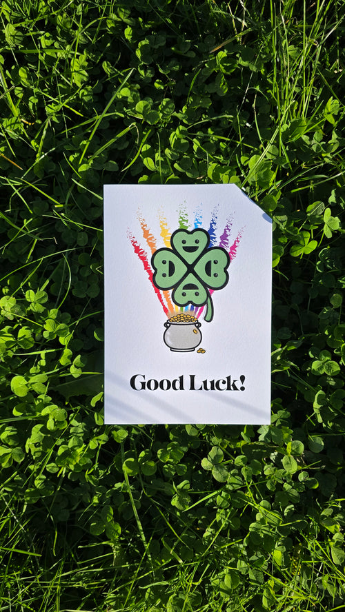 Card front with a smiling, cartoon four-leaf clover in the foreground, a rainbow in the background coming out of a pot of gold.
The text on the bottom reads 'Good Luck!', in bold black letters. The top right hand corner is cut off, for orientation purposes; a unique Dot & Type feature.
The image shows the card lay on a ground of clover.