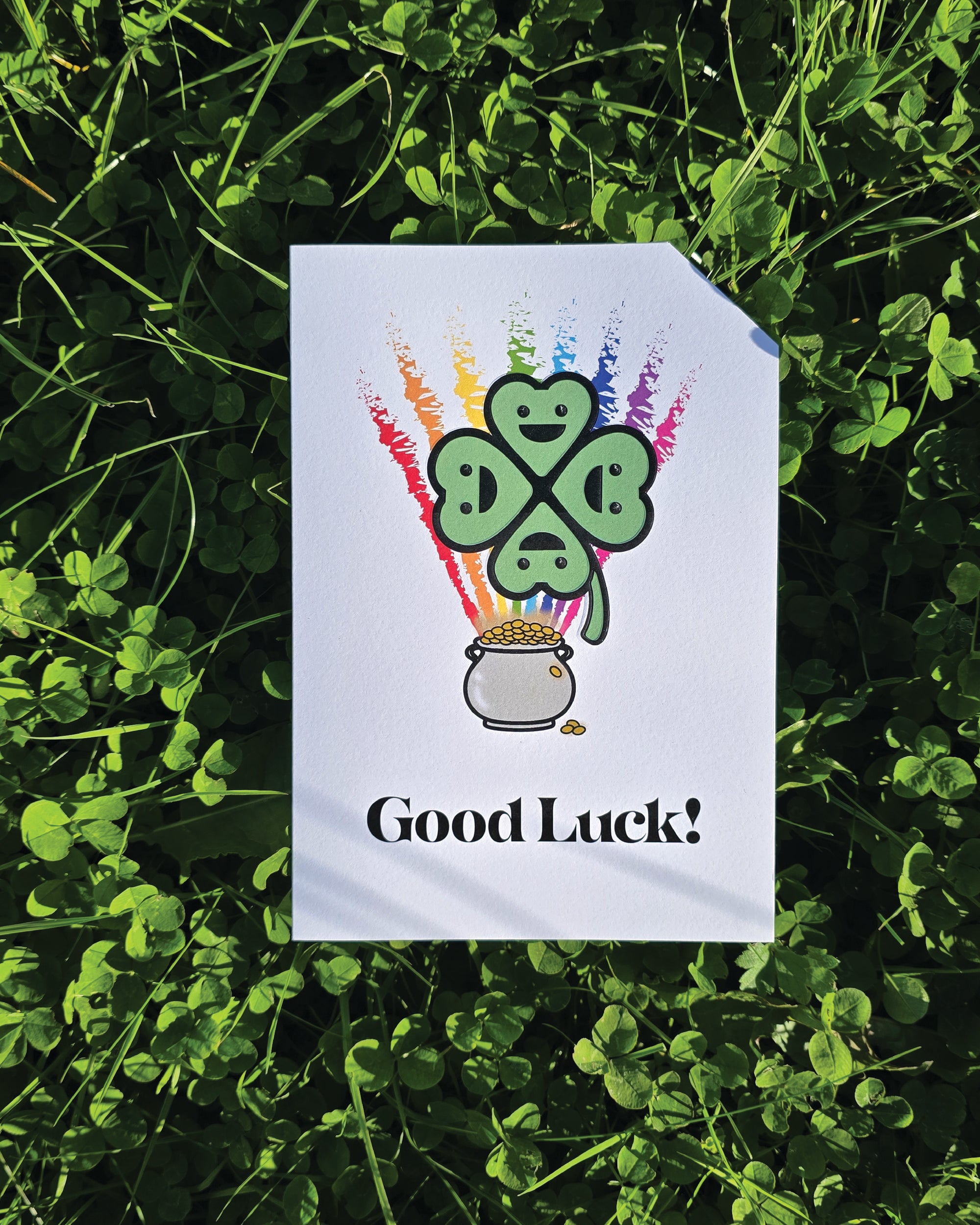 Card front with a smiling, cartoon four-leaf clover in the foreground, a rainbow in the background coming out of a pot of gold.
The text on the bottom reads 'Good Luck!', in bold black letters. The top right hand corner is cut off, for orientation purposes; a unique Dot & Type feature.
The image shows the card lay on a ground of clover.