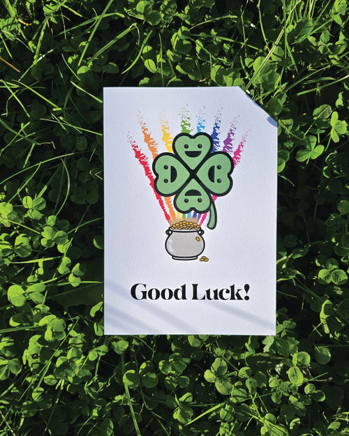Card front with a smiling, cartoon four-leaf clover in the foreground, a rainbow in the background coming out of a pot of gold.
The text on the bottom reads 'Good Luck!', in bold black letters. The top right hand corner is cut off, for orientation purposes; a unique Dot & Type feature.
The image shows the card lay on a ground of clover.