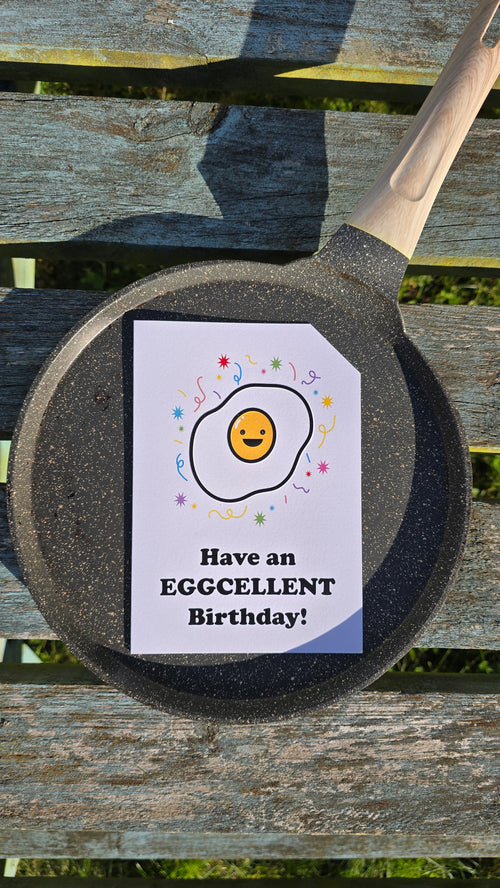 Card front with a smiling, cartoon fried egg with colourful confetti in the background.
The text on the bottom reads 'Have an EGGCELLENT Birthday!', in bold black letters. The top right hand corner is cut off, for orientation purposes; a unique Dot & Type feature.
The image is of the greetings card on top of a frying pan and it is placed on a wooden bench.
