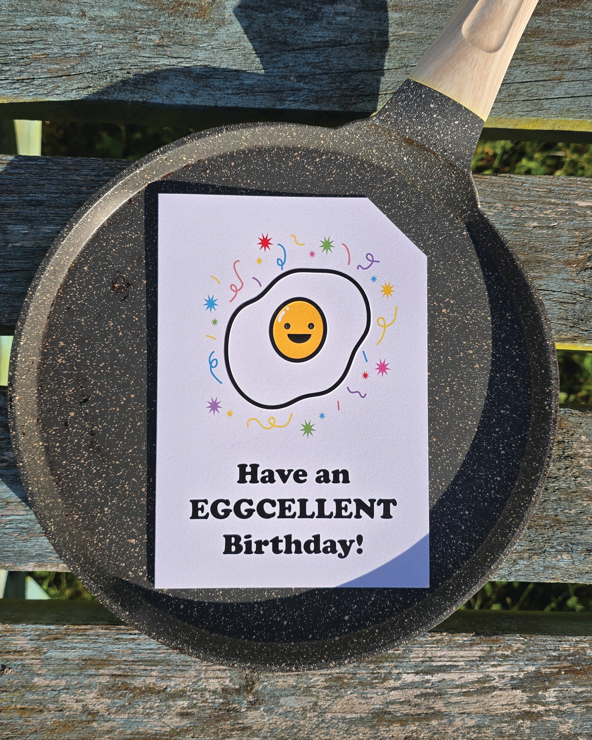 Card front with a smiling, cartoon fried egg with colourful confetti in the background.
The text on the bottom reads 'Have an EGGCELLENT Birthday!', in bold black letters. The top right hand corner is cut off, for orientation purposes; a unique Dot & Type feature.
The image is of the greetings card on top of a frying pan and it is placed on a wooden bench.