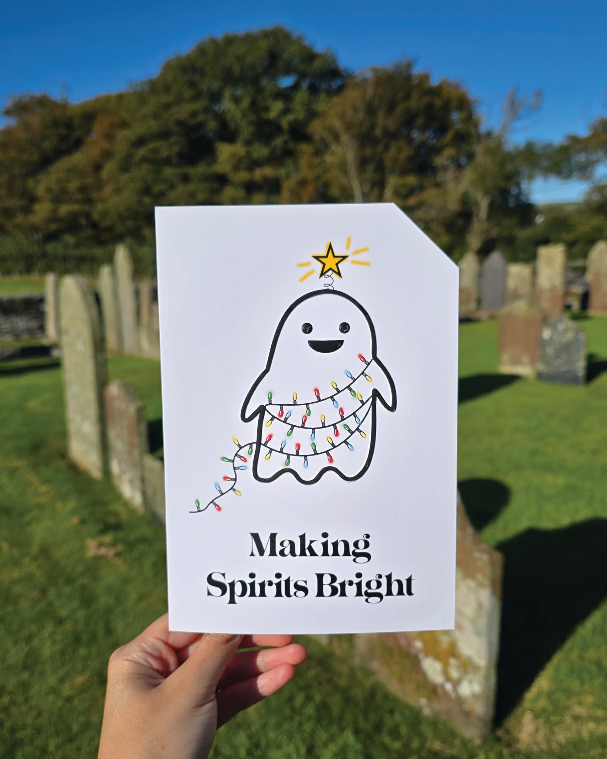 Card front with a smiling ghost wrapped in red, yellow, green and blue, Christmas tree lights. The ghost has a glowing yellow star on its head.
The text on the bottom reads 'Making Spirits Bright', in bold black letters. The top right hand corner is cut off, for orientation purposes; a unique Dot & Type feature.
The image was taken in a graveyard, the card is in the foreground and there are old graves in the background. The sky is bright blue and cloudless.