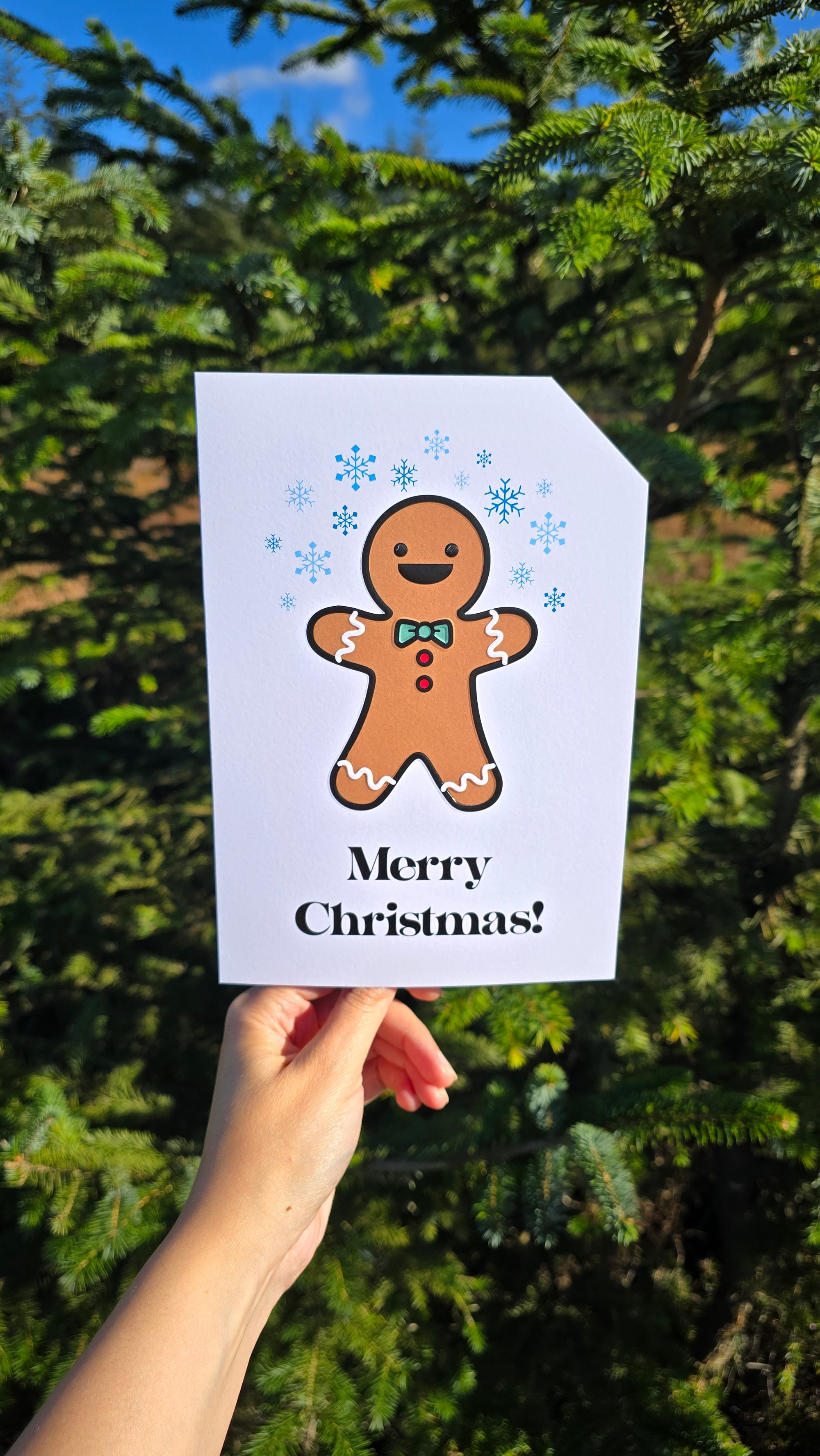 Card front with a smiling gingerbread person in the foreground. They are wearing a green bow-tie with red buttons. White icing waves are on their arms and legs. There are various blue snowflakes in the background.
The text on the bottom reads 'Merry Christmas!', in bold black letters. The top right hand corner is cut off, for orientation purposes; a unique Dot & Type feature.
The image is of the card taken in front of a outdoor Christmas tree.