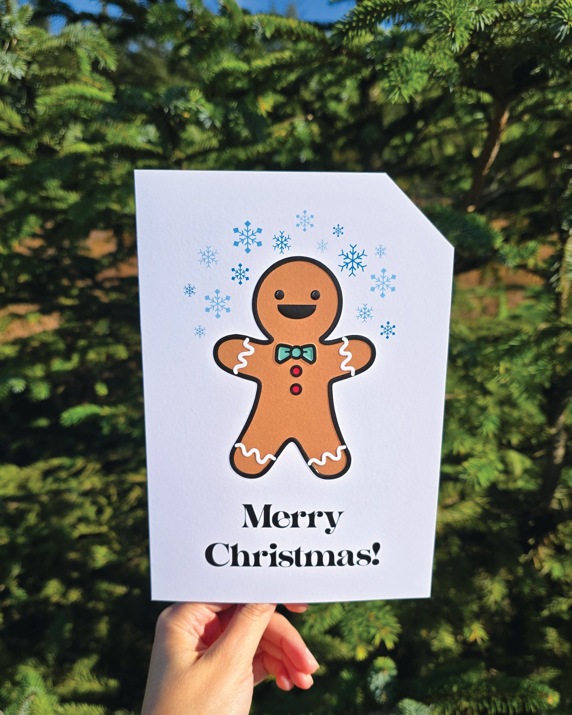 Card front with a smiling gingerbread person in the foreground. They are wearing a green bow-tie with red buttons. White icing waves are on their arms and legs. There are various blue snowflakes in the background.
The text on the bottom reads 'Merry Christmas!', in bold black letters. The top right hand corner is cut off, for orientation purposes; a unique Dot & Type feature.
The image is of the card taken in front of a outdoor Christmas tree.