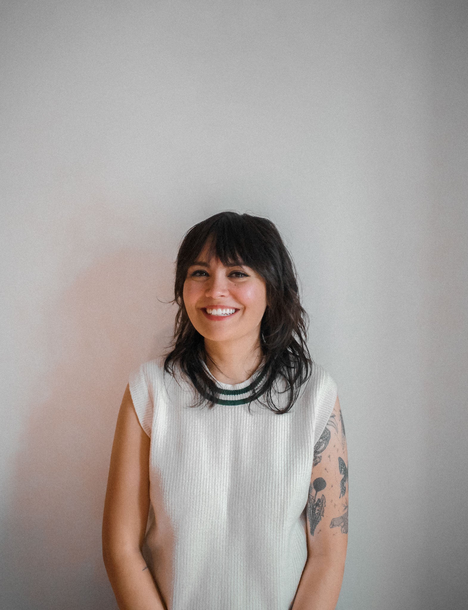 Woman with dark, layered hair and half-sleeve tattoo on left arm. Wearing a light-colored sleeveless top against a plain background. She is smiling whilst looking at the camera.