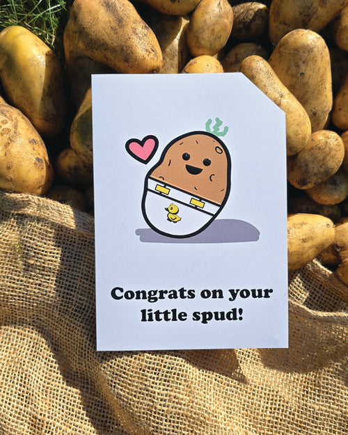 Card front with a smiling, cartoon potato character wearing a nappy/diaper with a duck on it. There is a small pink heart next to the potato. The text on the bottom reads 'Congrats on your little spud!', in bold black letters. The top right hand corner is cut off, for orientation purposes. A unique Dot & Type feature.
The image is of the card lay on a hessian sack which has potatoes falling out of it.