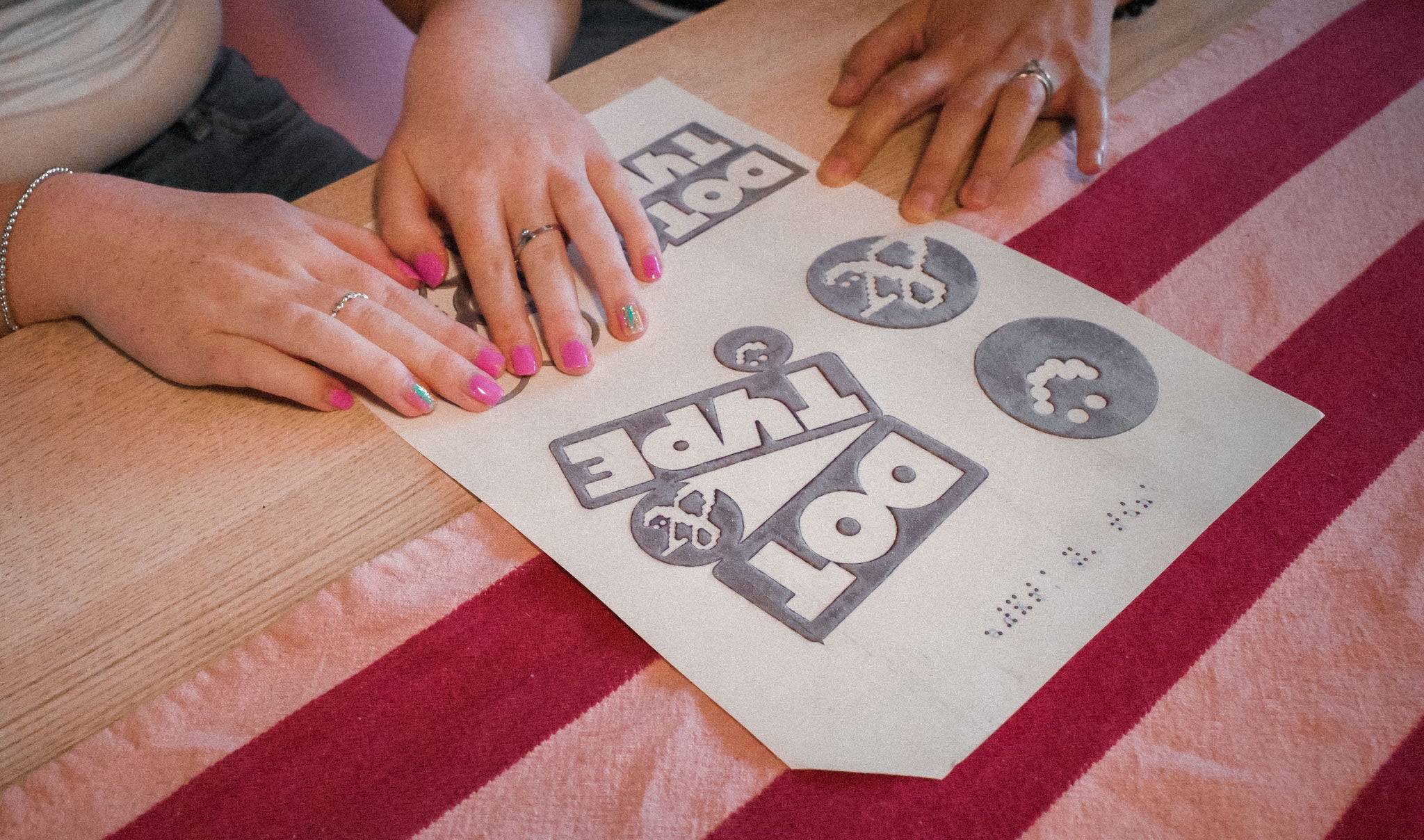 Close up hands of a young blind lady, reading the tactile image with the logo DOT & TYPE and different icons associated, all on tactile image paper. She is being supported by someone else holding the paper down. The young lady has pink nail varnish on. Striped fabric on the table, in the background.
