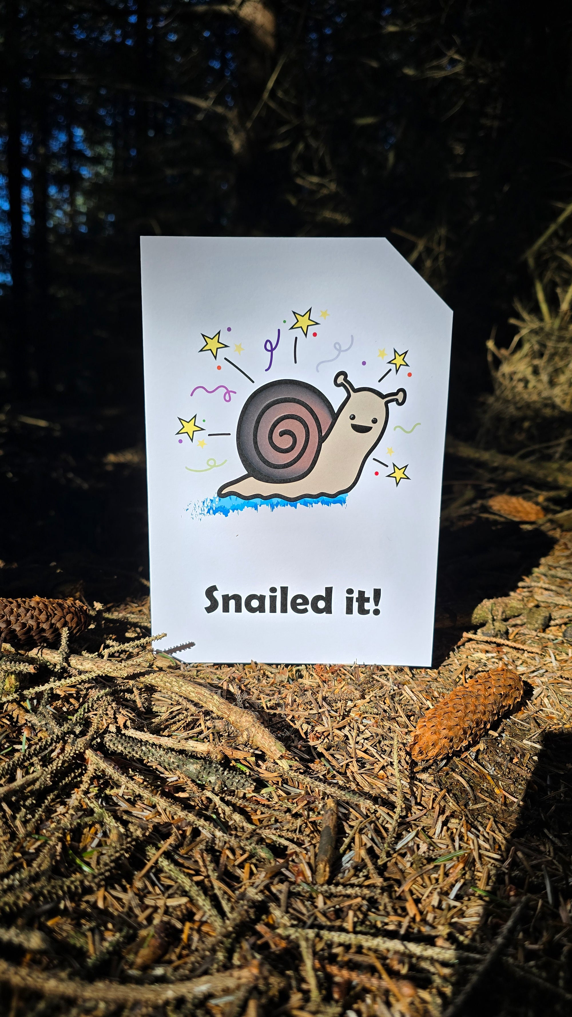 Card front with a smiling snail with a blue snail trail in the foreground. There are stars and colourful confetti in the background.
The text on the bottom reads 'Snailed it!', in bold black letters. The top right hand corner is cut off, for orientation purposes; a unique Dot & Type feature.
The image is of the card p[laced on the forest floor. There are twigs and pine cones lay on the floor.