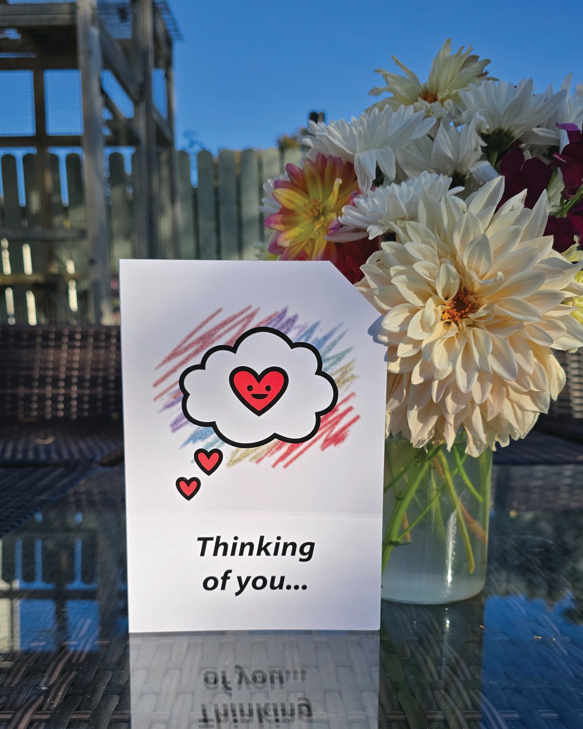 Card front with a 'thinking cloud' with a smiling red heart in the centre, in the foreground. Colourful aray of scribbles in the background.
The text on the bottom reads 'Thinking of you...', in bold black letters. The top right hand corner is cut off, for orientation purposes; a unique Dot & Type feature.
The image shows the card on a table with a small vase of flowers next to it. There are large homegrown dahlias in the vase.