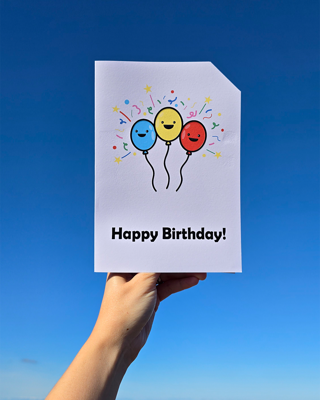 Card front with 3 smiling, cartoon balloons, with bright confetti type graphics in the background. Left balloon is pale blue, middle balloon is yellow and the right balloon is red.
The text on the bottom reads 'Happy Birthday!', in bold black letters. The top right hand corner is cut off, for orientation purposes; a unique Dot & Type feature.
The image is of the card being held in front of a bright blue, cloudless sky.