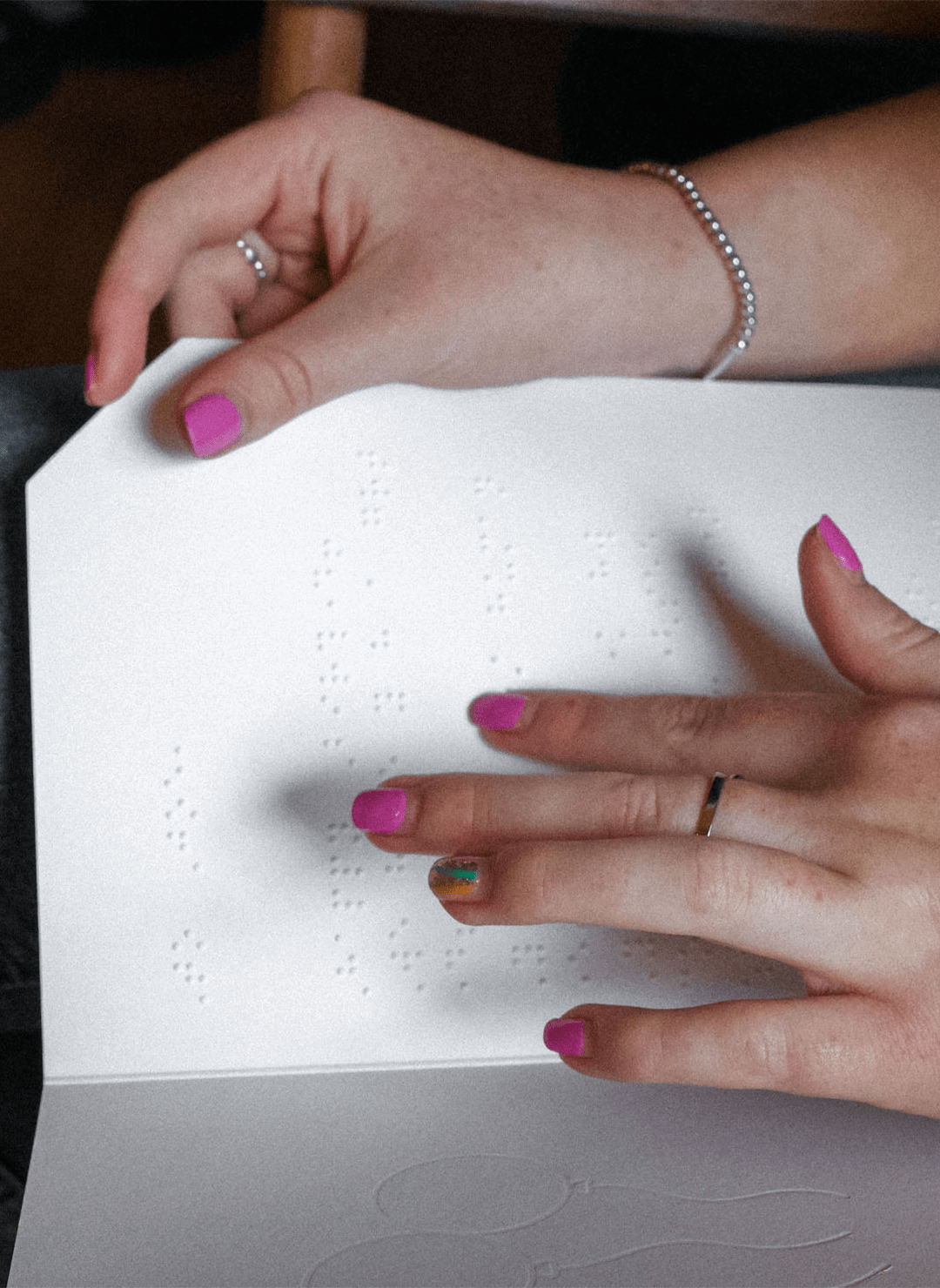 A close up of a young lady's hands reading braille in a braille greetings card. She has pink nail polish on and a small silver beaded bracelet on her right arm.